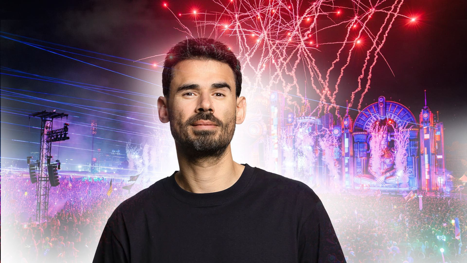 Afrojack in a black shirt faces the camera with a vibrant festival stage and fireworks behind him.