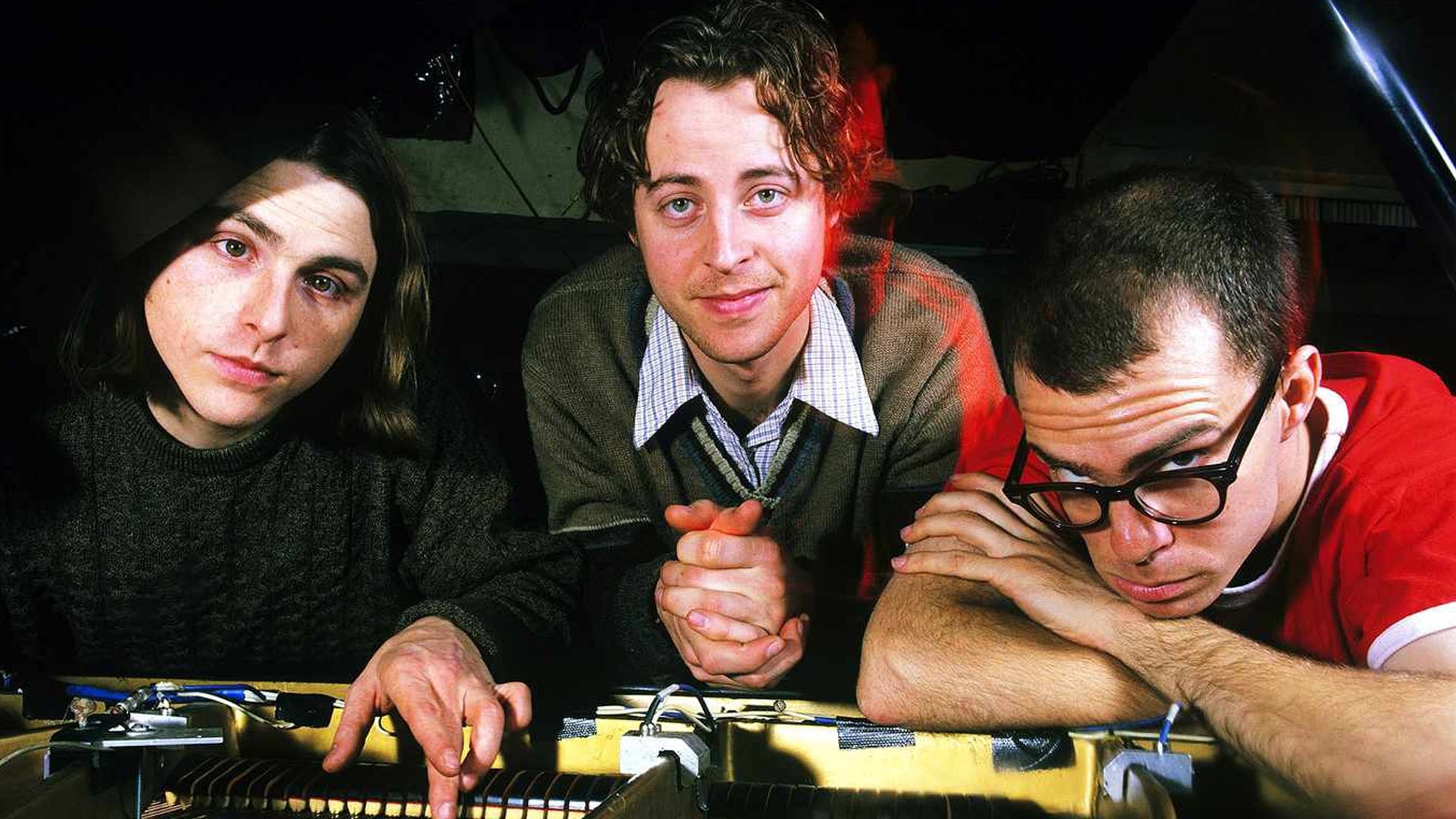 Ben Folds Five autour d'un piano ouvert dans une pièce sombre : l'un en pull foncé, l'un en cardigan et l'un en rouge avec des lunettes.