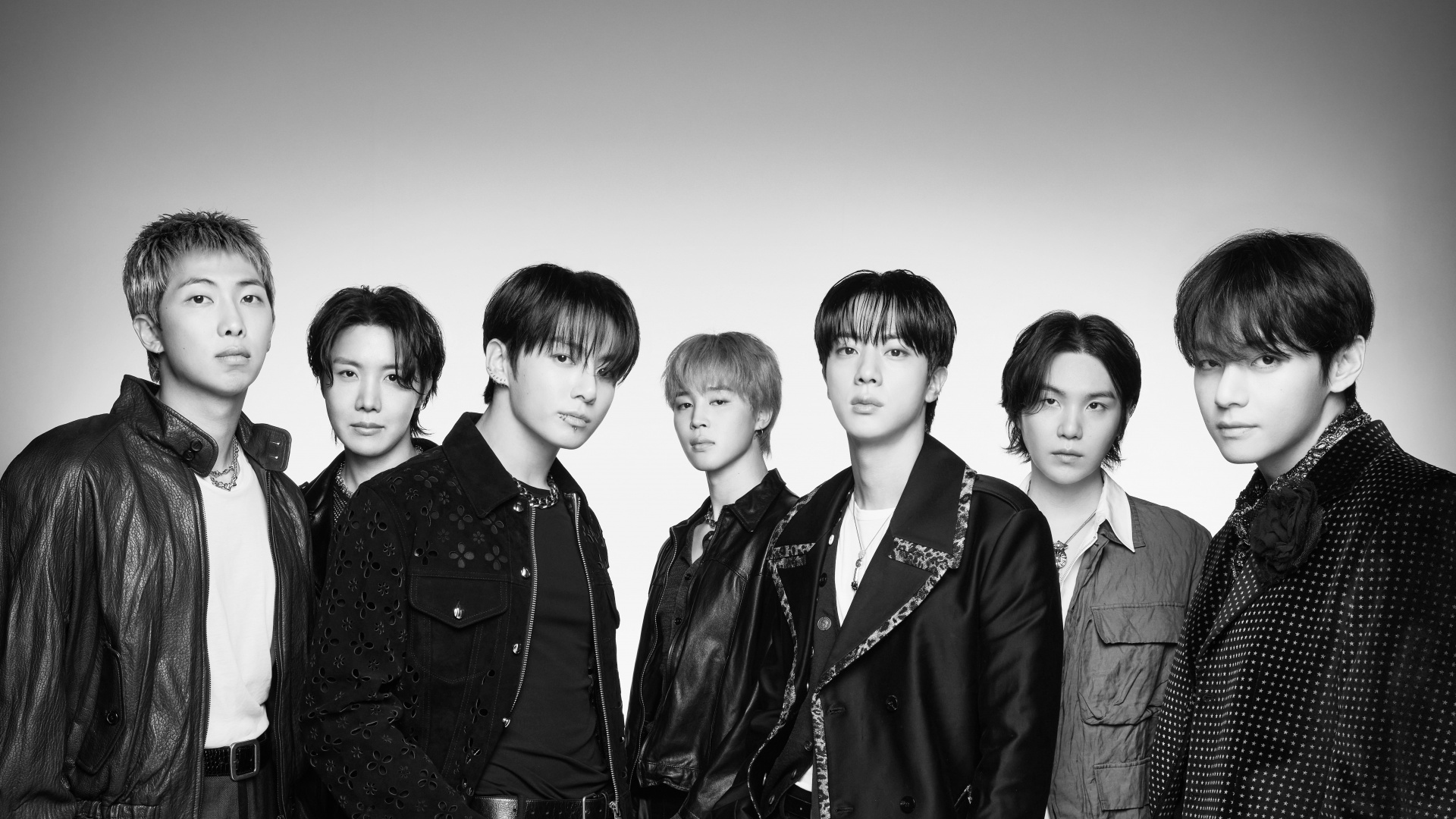 Black-and-white group portrait of all seven BTS members in dark leather and formal jackets against a plain studio background