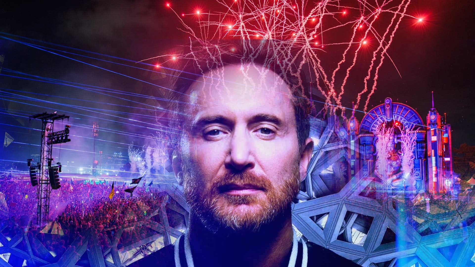 David Guetta at an electric festival stage with neon lights, crowds, and pyrotechnics overhead.