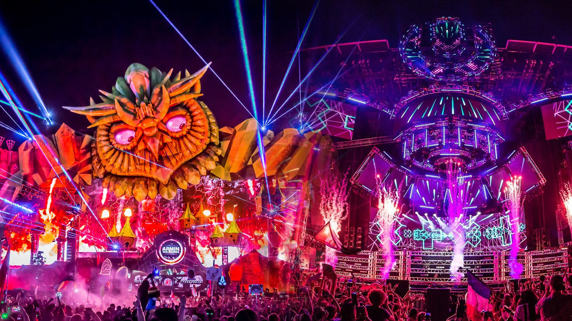 Scène de festival de nuit avec une sculpture géante de hibou orange, des faisceaux laser, de la pyrotechnie et une foule dense aux bras levés.