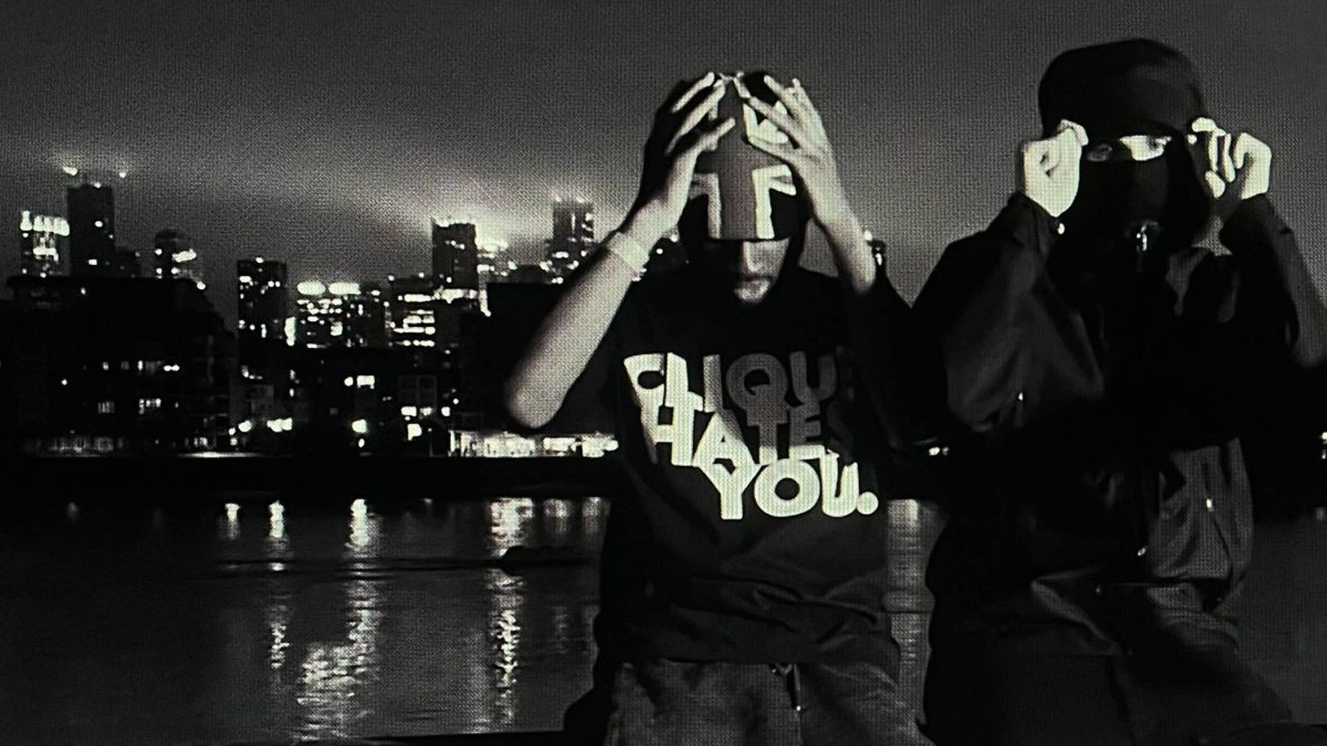 Two people in dark clothing cover their faces at night in black and white, with a glowing city skyline over water behind them.