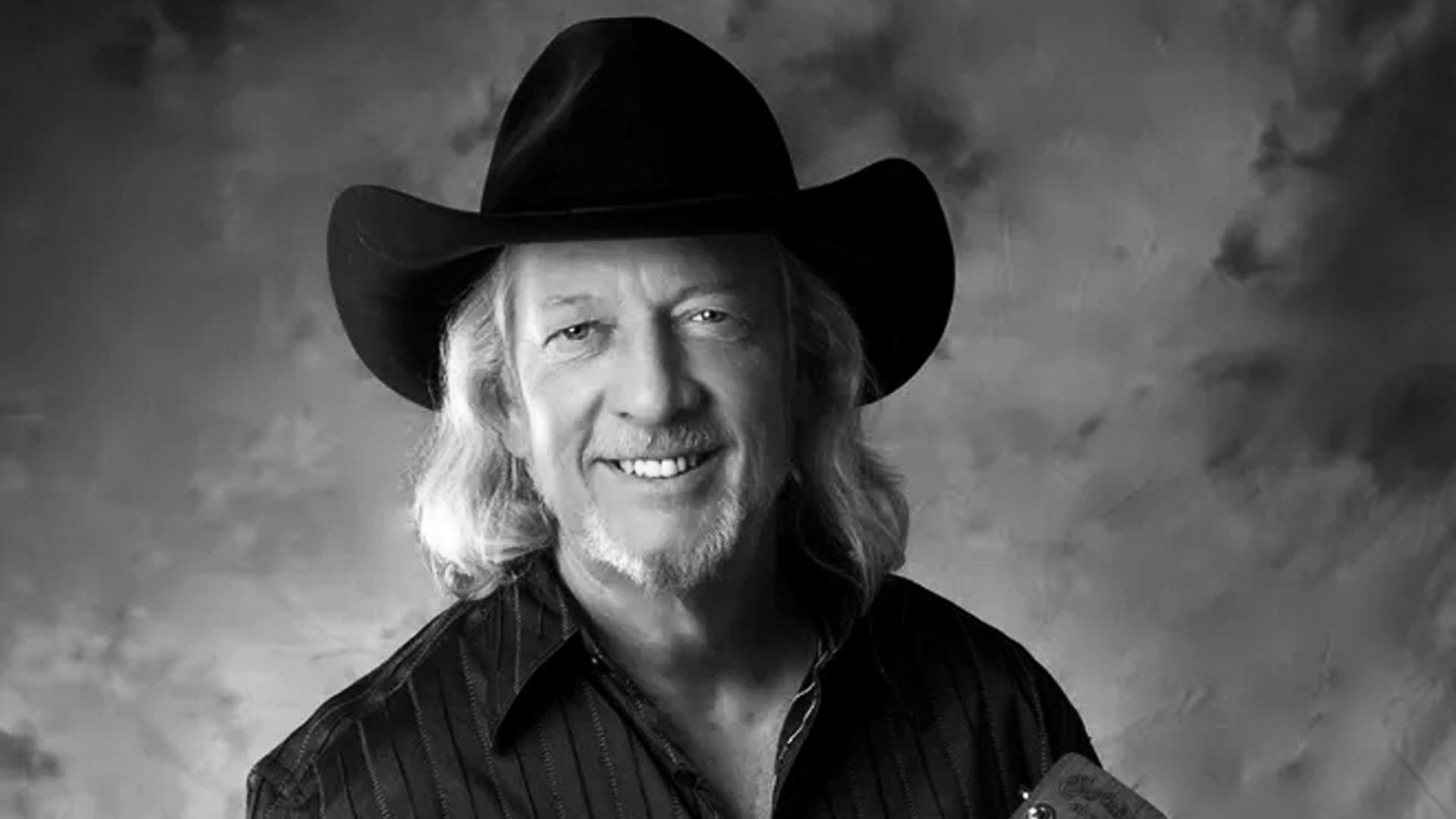 Black-and-white portrait of John Anderson in a black cowboy hat and dark striped shirt, with long gray hair and a goatee.