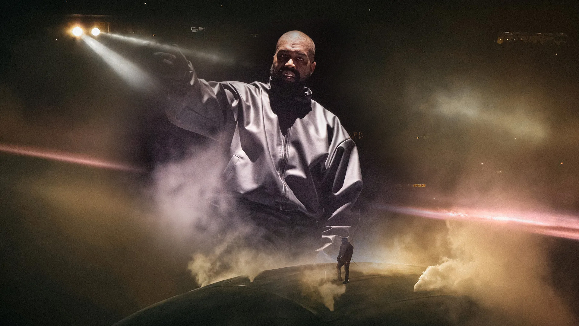 A giant Kanye West towers over the globe-shaped stage of the Ye Live Tour, wreathed in smoke and spotlights, with a tiny figure standing on the globe below