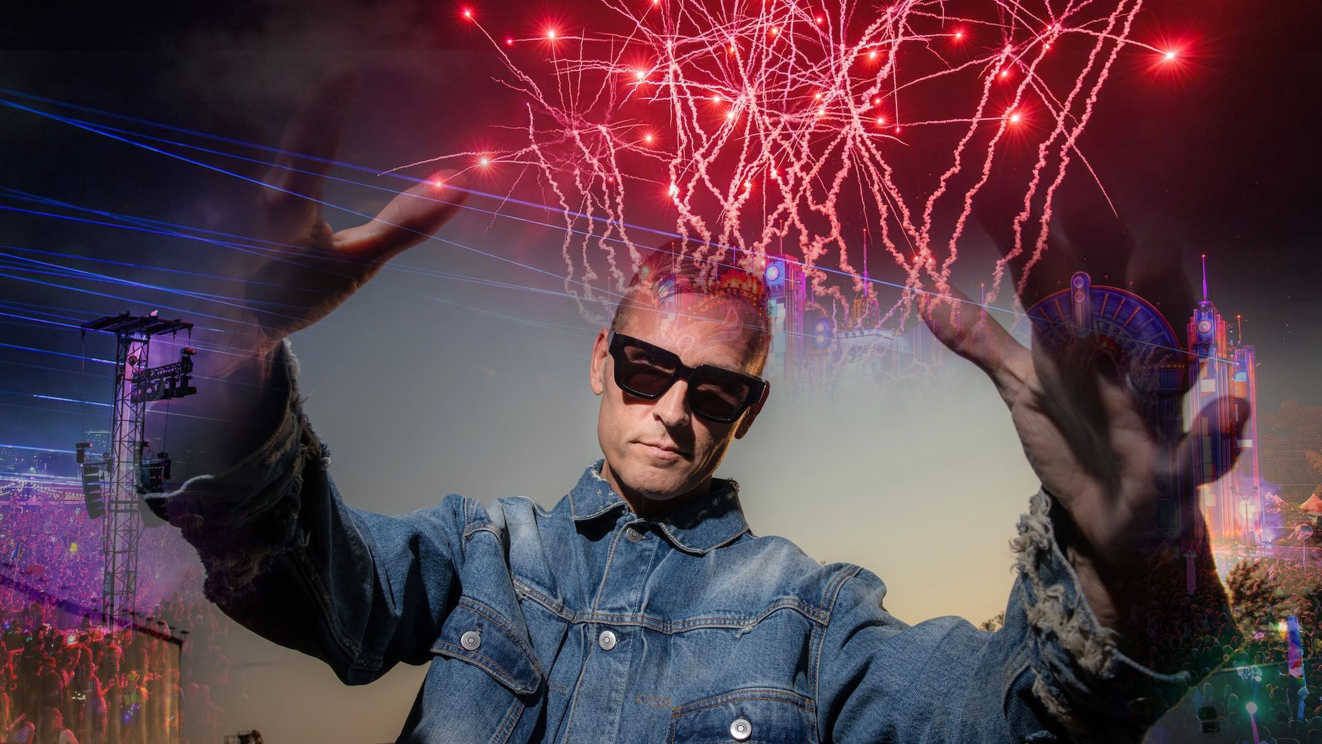Kaskade wearing dark sunglasses and denim jacket with hands raised beneath vibrant red fireworks and stage lights.