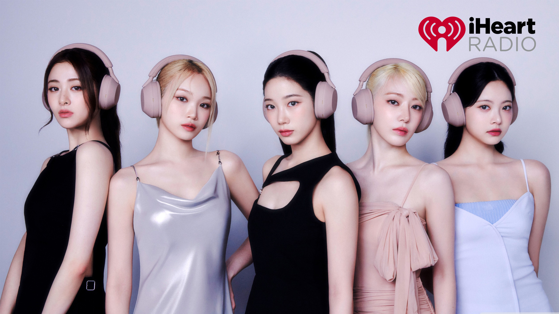 LE SSERAFIM's five members posing together in profile, wearing matching blush-pink over-ear headphones and a mix of black, silver, and pastel dresses, against a plain grey studio background with the iHeartRadio logo in the corner