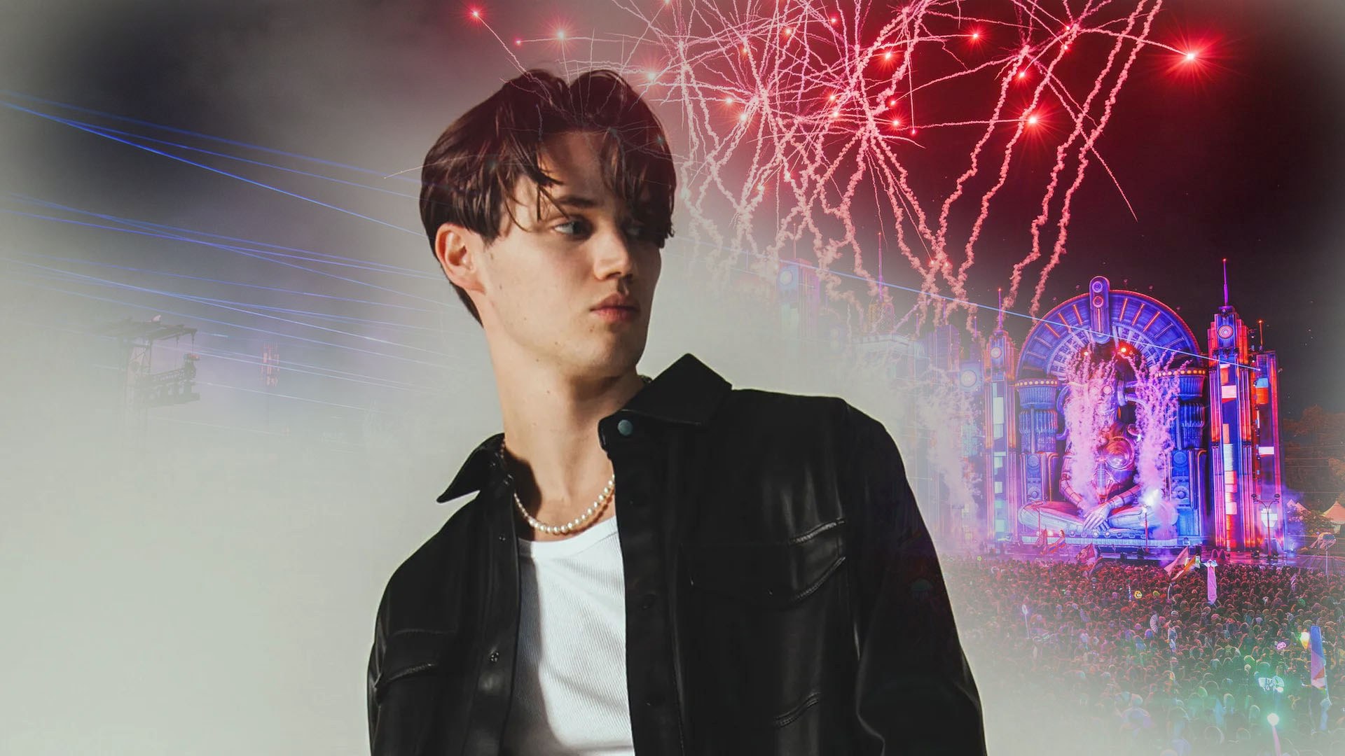 Mau P in a black jacket with a white shirt and chain necklace, standing behind a festival venue with red and blue lights