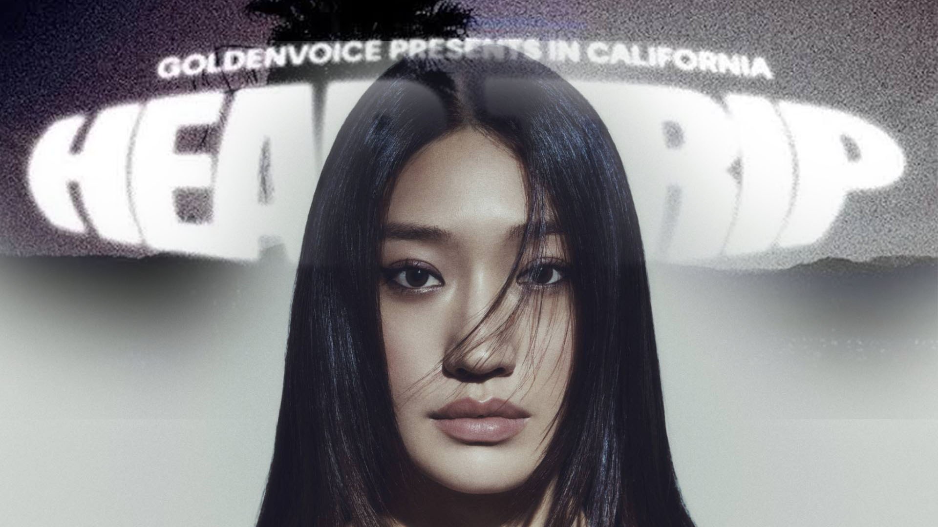 Peggy Gou на фестивальной сцене с длинными тёмными волосами, смотрит прямо в камеру на фоне светящихся вывесок.