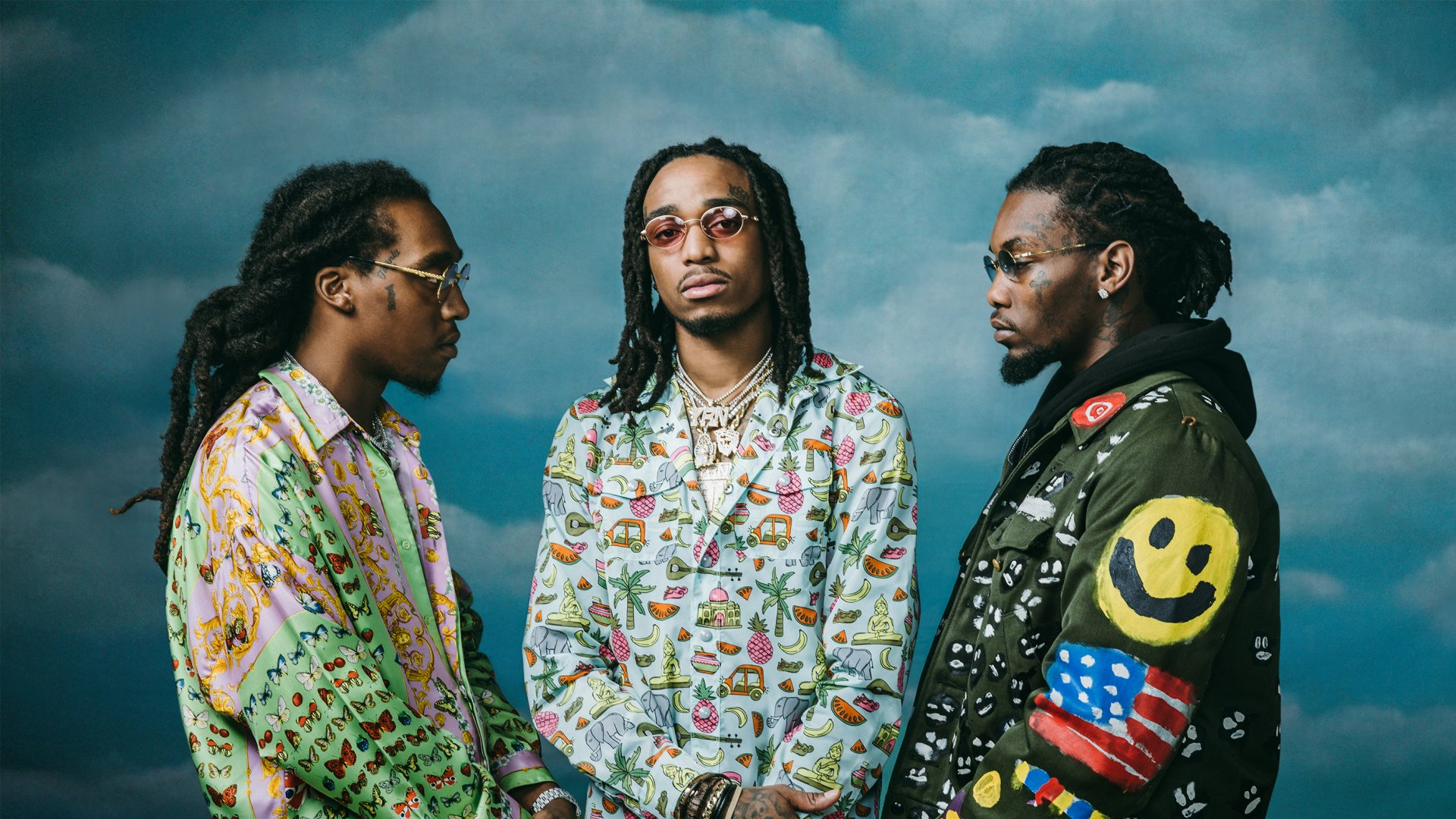 An archival photo of Migos — Quavo, Offset, and Takeoff — standing in profile facing each other against a cloudy sky, each wearing colorful printed outfits
