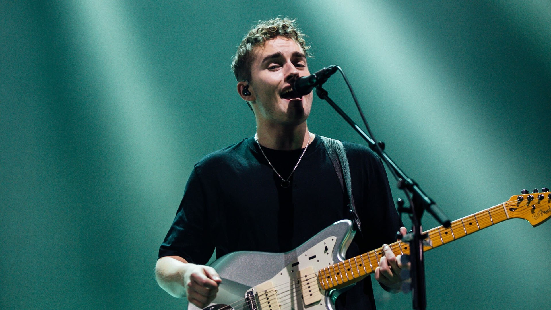 Sam Fender in a black t-shirt sings into a microphone while playing a silver electric guitar under teal lights.