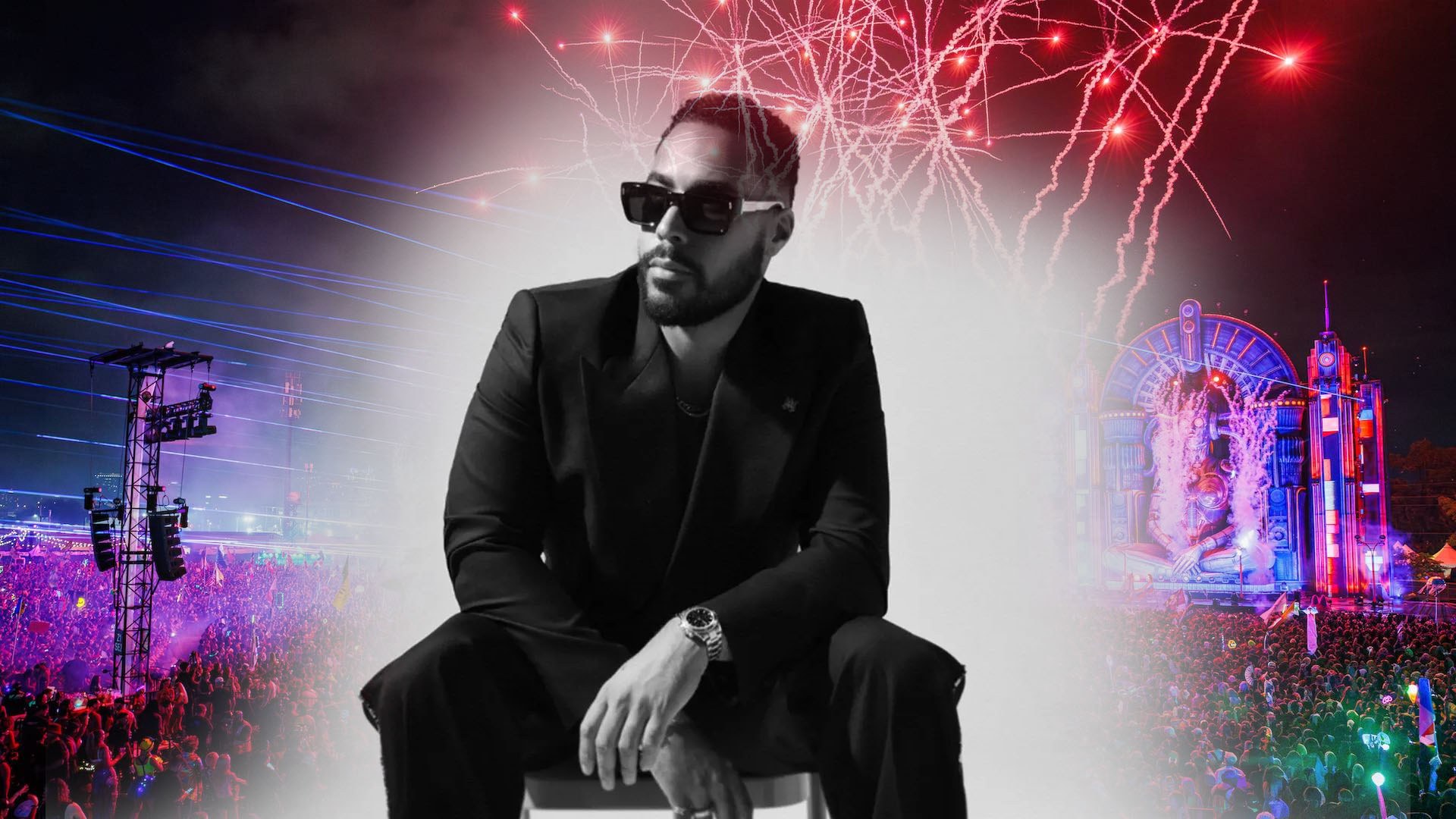 Man in a black suit and sunglasses seated on a stool in black-and-white, framed by a festival crowd and red fireworks.